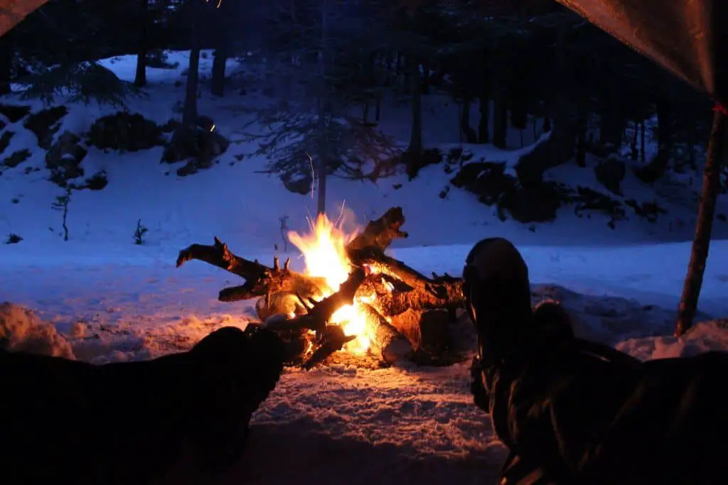 two people lying in a tent wearing the best thermal underwear for extreme cold warming their feet at a fire in the snow