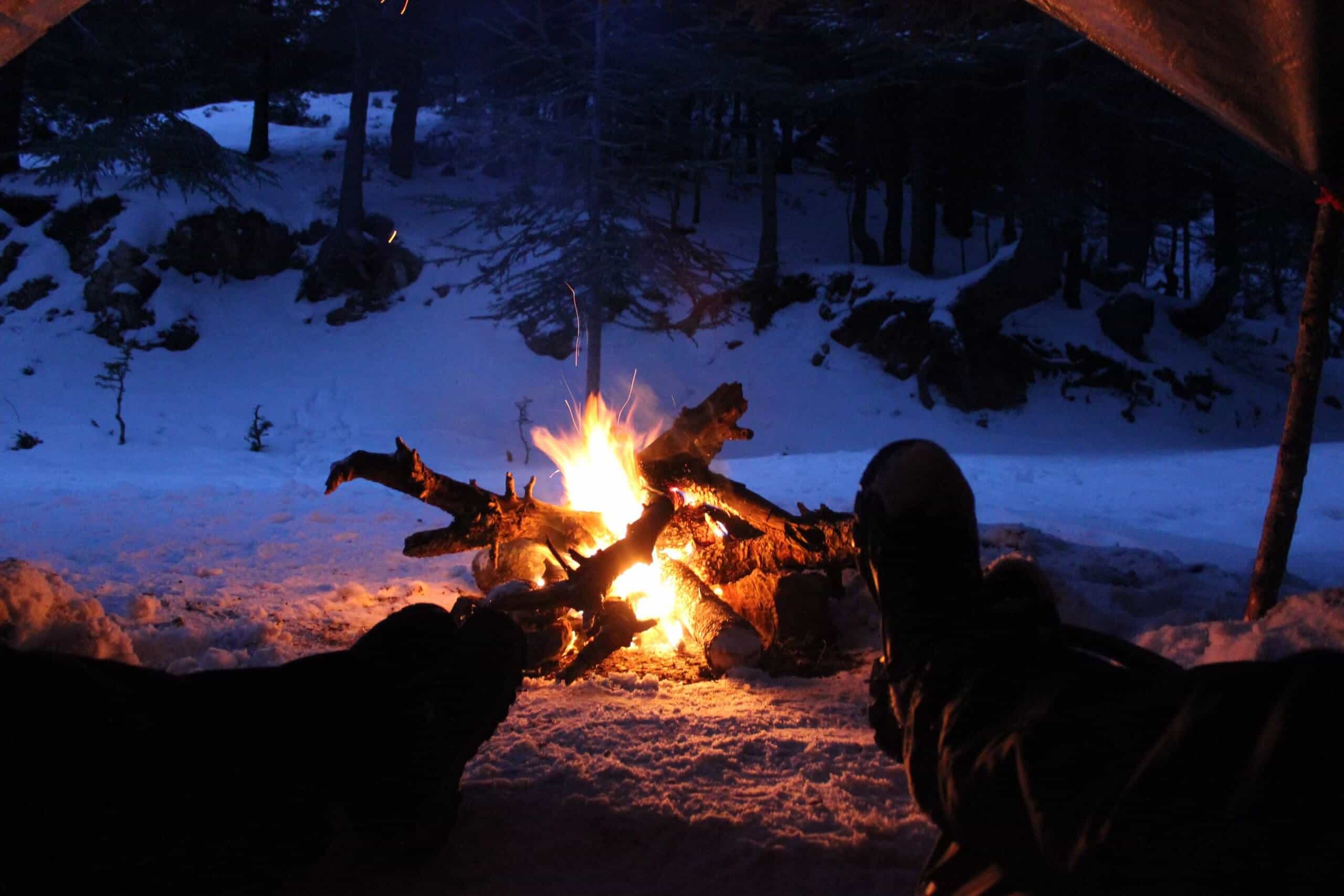 two people lying in a tent wearing the best thermal underwear for extreme cold warming their feet at a fire in the snow