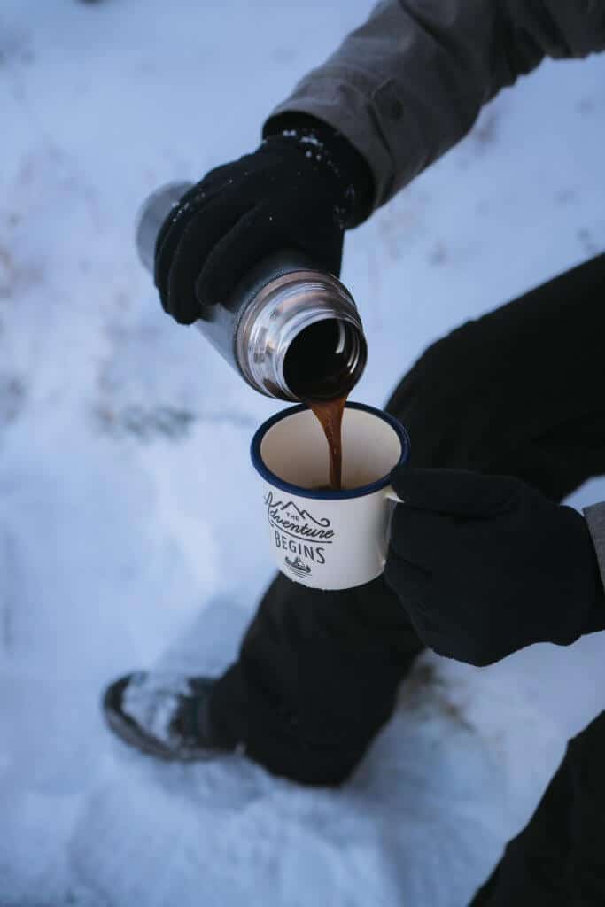 man pouring coffee in a mug while sitting in the snow wearing ice fishing gloves