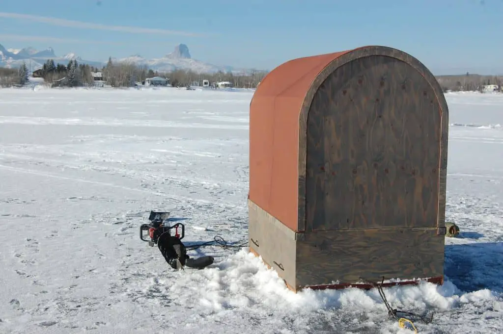 One of the best battery powered ice augers lying in the snow on a frozen lake next to an ice shed
