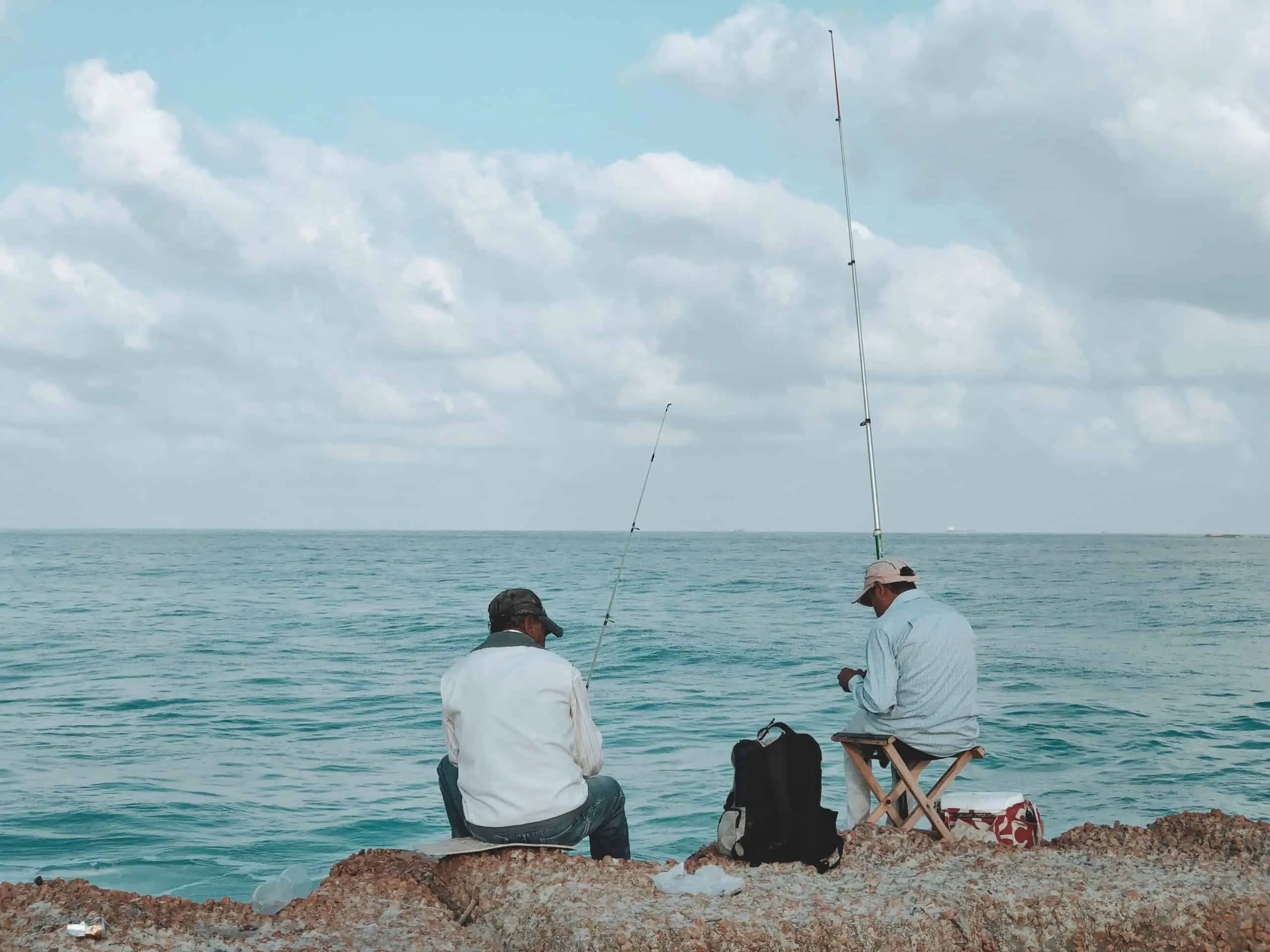 Fishing requires patience: two men sitting on rocks in front of the ocean holding rods waiting for fish to bite