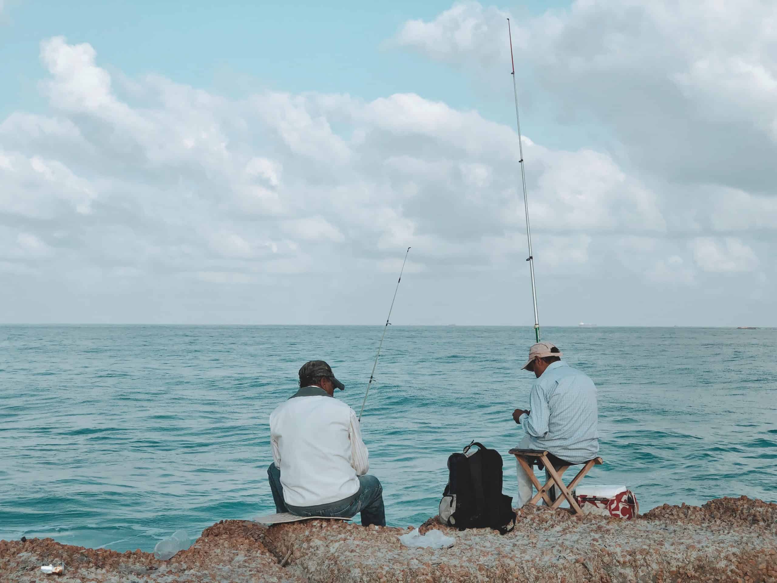 Fishing requires patience: two men sitting on rocks in front of the ocean holding rods waiting for fish to bite