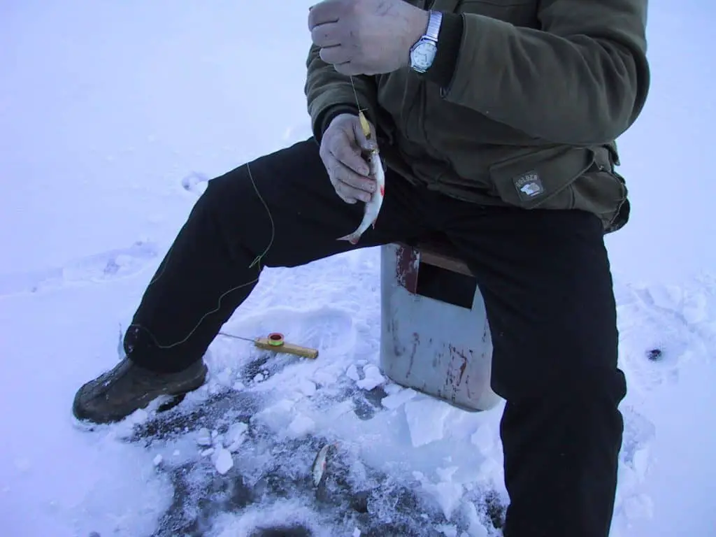 Man wearing some of the best ice fishing boots reeling in a catch from a drilled hole on a frozen lake