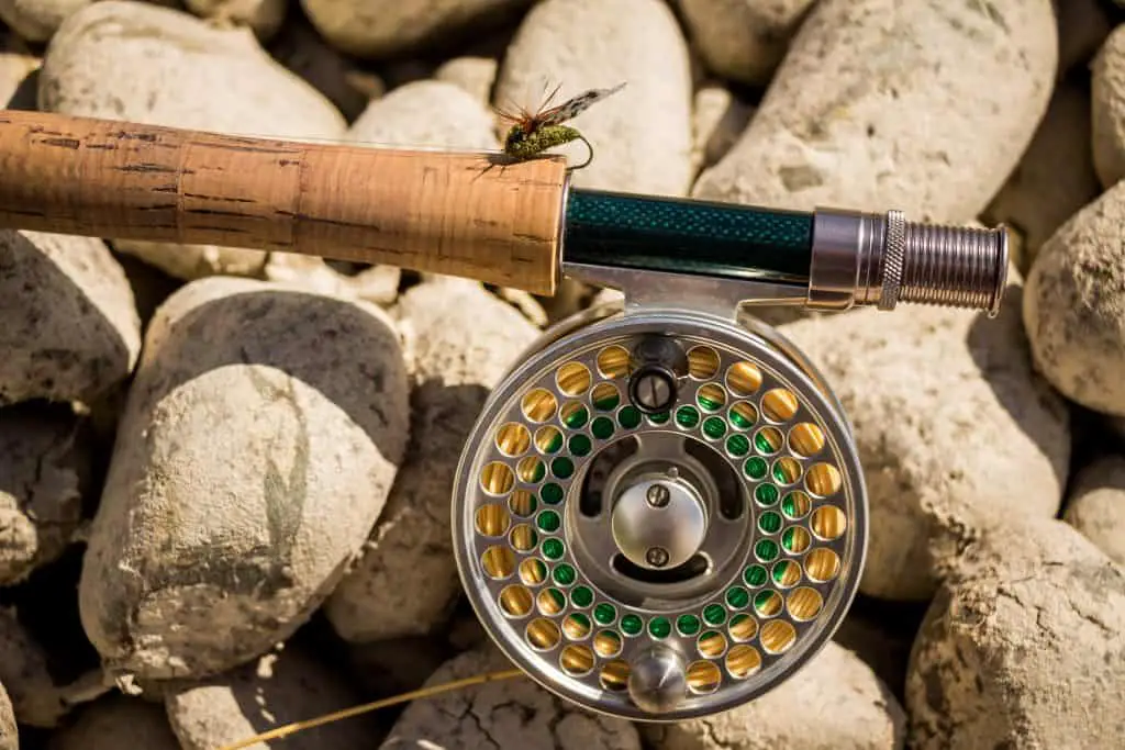 A close up of a trout fly rod, reel and line on rocks, with an artificial cicada fly attached to the cork as a symbol for the best fly fishing brands